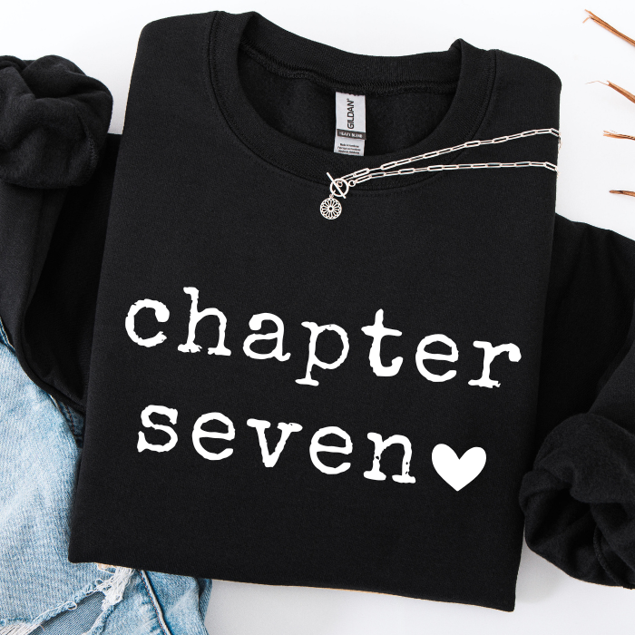 A black crewneck sweatshirt with 'chapter seven' and a heart printed in white on the front, styled flat-lay with a silver chain necklace and light blue denim.
