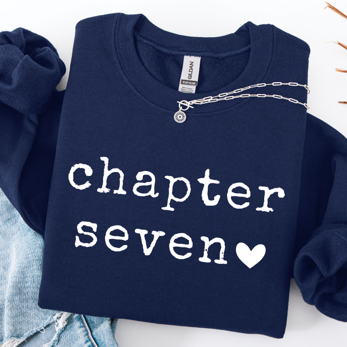 A navy blue sweatshirt with 'chapter seven' and a heart printed in white across the front, styled flat-lay with light blue denim and a silver chain necklace on a white background.