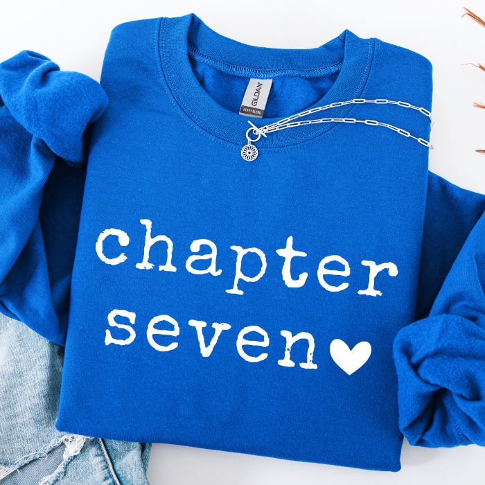 A folded royal blue sweatshirt with rolled sleeves, featuring the words 'chapter seven' in white text and a white heart. A silver chain necklace is draped across the collar, and light-wash denim jeans are partially visible at the bottom left.