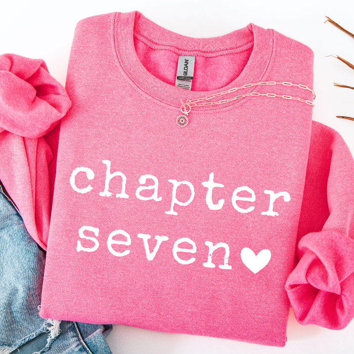 A folded pink long-sleeve sweatshirt with 'chapter seven' printed in white lowercase letters and a white heart, styled with rolled sleeves and a gold chain necklace on a white background.