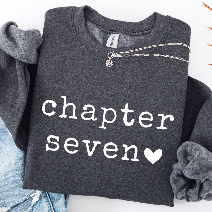 A dark heather gray sweatshirt with 'chapter seven' printed in white lowercase letters and a white heart, styled flat-lay with a silver chain necklace, blue jeans, and a gray scrunchie on a white background.