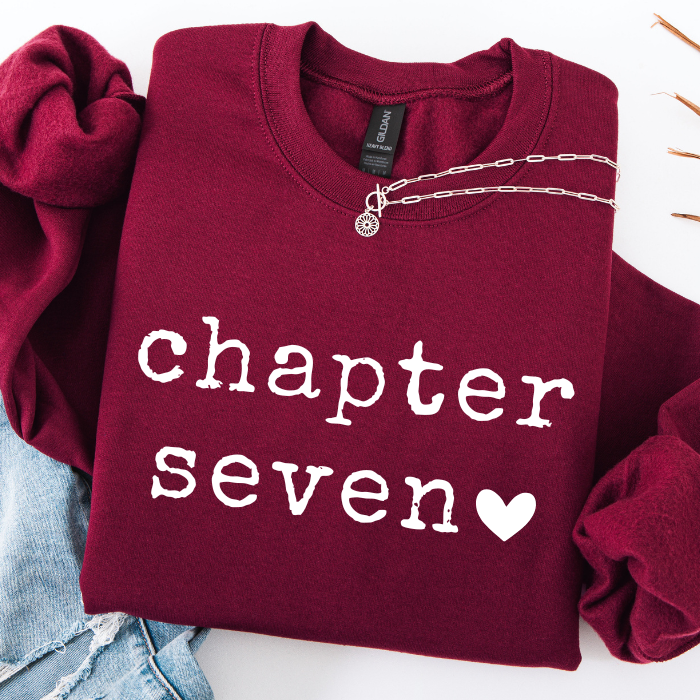 A folded burgundy sweatshirt with 'chapter seven' printed in white across the front, styled with a silver chain necklace and light blue jeans on a white background.