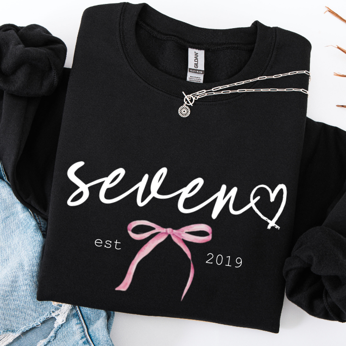 A folded black long-sleeve sweatshirt with 'seven est 2019' and a pink bow graphic on the front, styled with a silver necklace and light blue denim on a white background.