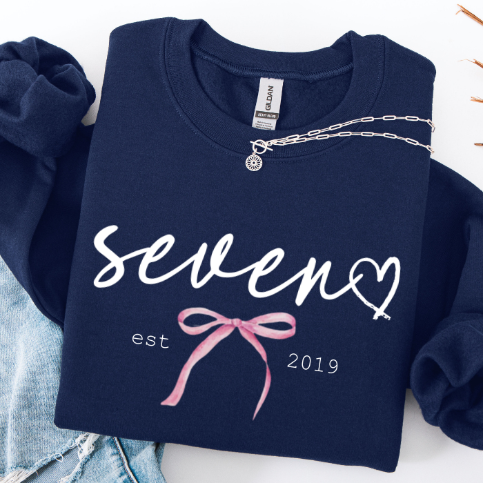 A navy blue sweatshirt laid flat on a white surface, featuring the word 'seven' in white script with a heart shape formed by the final letter, plus 'est 2019' and a pink bow graphic. A silver chain necklace is draped across the chest, and light blue denim is partially visible at the lower left.