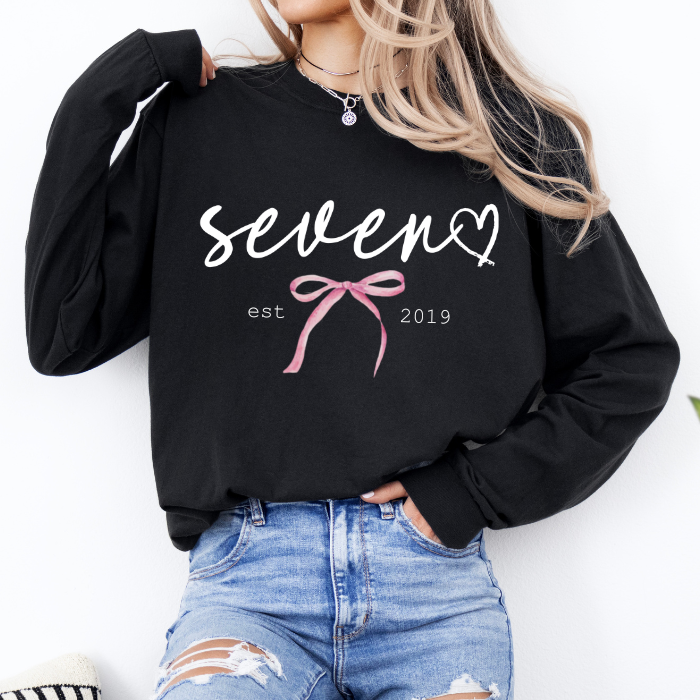 A woman wearing a black long-sleeve sweatshirt with 'seven est 2019' and a pink bow graphic on the front, paired with distressed blue jeans.