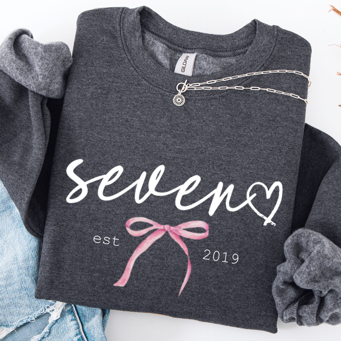 A folded dark heather gray sweatshirt with a white script 'seven' and a pink bow graphic, styled with a silver necklace and light blue jeans on a white background.