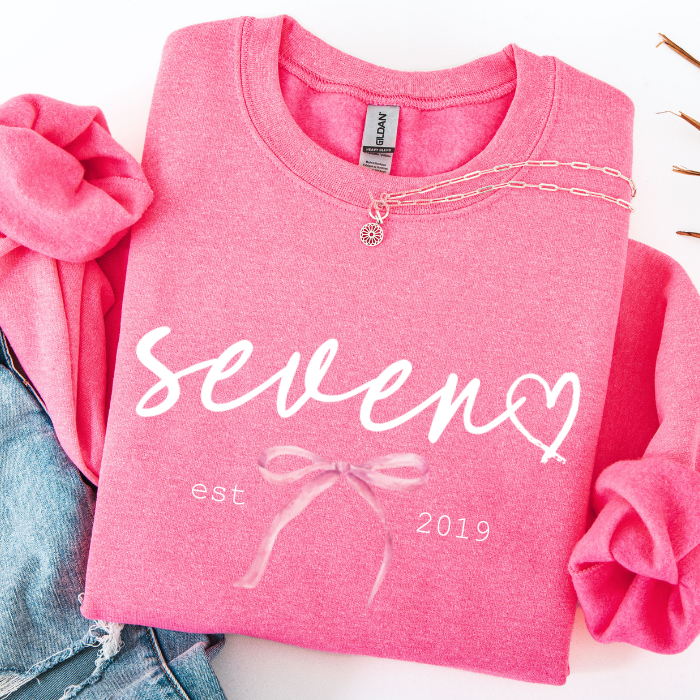 A folded pink sweatshirt with rolled sleeves, featuring the word 'seven' in white script with a heart shape formed by the 'n', plus 'est. 2019' and a bow graphic.