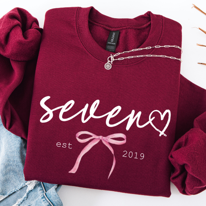 A folded burgundy sweatshirt with white script text reading 'seven' and 'est 2019', plus a pink bow graphic, styled on a white background with a silver necklace draped across the chest and light blue jeans partially visible at the lower left.