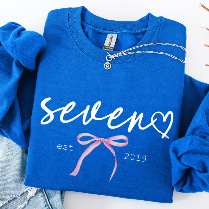 A folded royal blue sweatshirt with rolled sleeves, featuring a white script 'seven' with a heart, 'est 2019' text, and a pink ribbon graphic. A silver chain necklace is draped over the collar, and light blue denim jeans are partially visible at the bottom left.