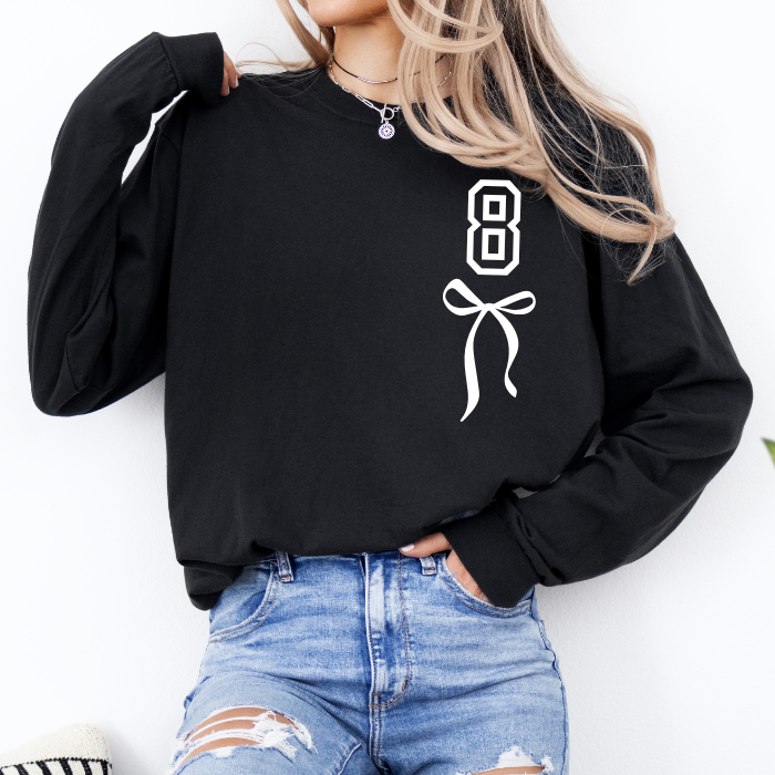 A woman wearing a black long-sleeve sweatshirt with a white graphic print of the number 8 and a ribbon bow on the front, paired with distressed blue jeans.