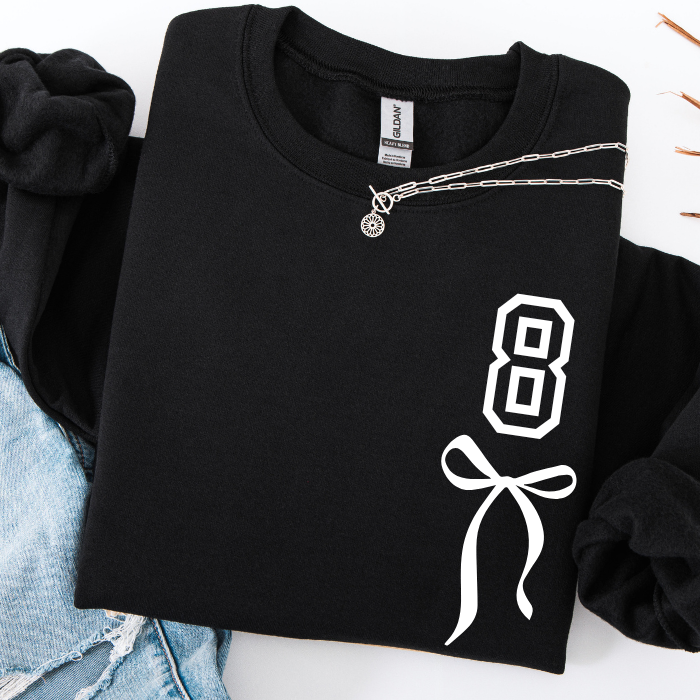 A folded black long-sleeve sweatshirt with a white graphic print of the number 8 and a bow on the front, styled with a silver chain necklace and light blue denim shorts on a white background.