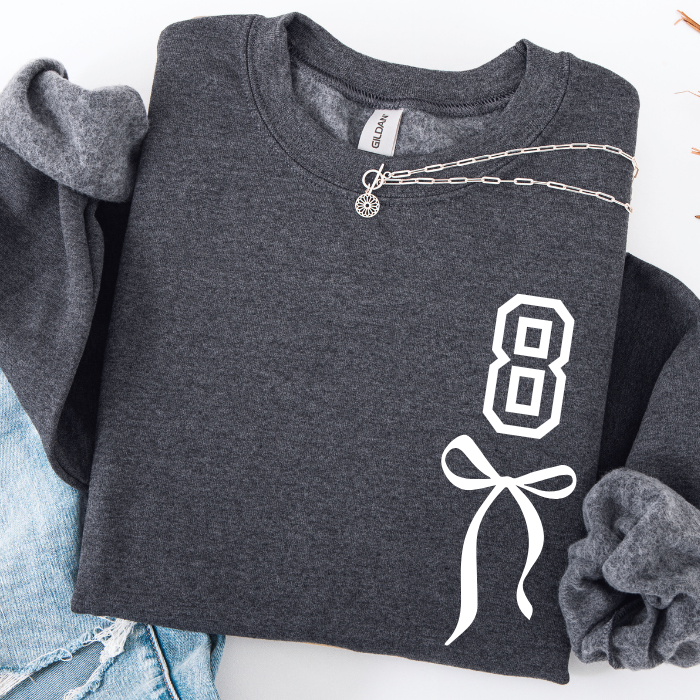 A dark gray sweatshirt folded flat on a white background, featuring a white graphic of the number 8 with a ribbon bow on the front. A silver chain necklace is draped across the chest, and a pair of light blue jeans is partially visible on the left.