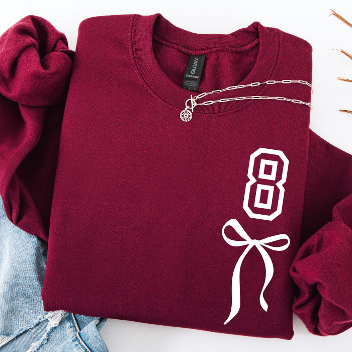 A folded burgundy crewneck sweatshirt with a white graphic print of the number 8 and a bow on the front, styled with rolled sleeves and a silver chain necklace on a white background.