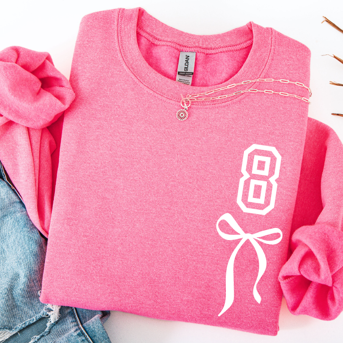 A folded pink crewneck sweatshirt with a white graphic of the number 6 and a bow on the front, styled with rolled sleeves and a gold chain necklace draped across the collar. A pair of blue jeans is partially visible on the left.