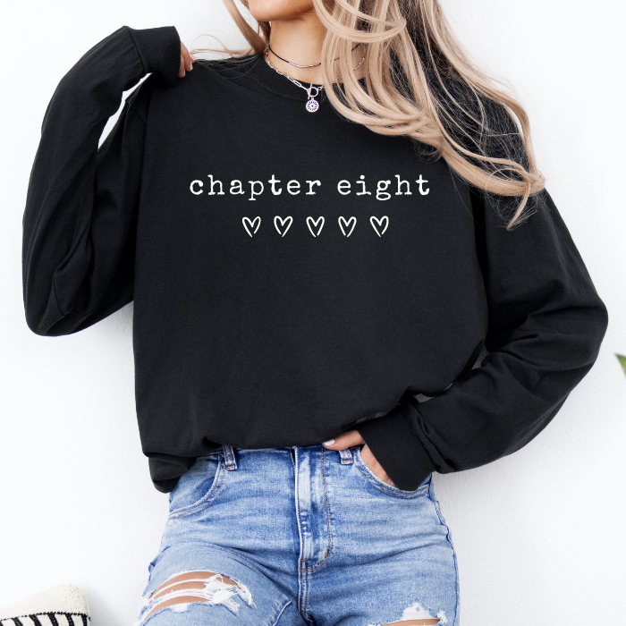 A woman wearing a black long-sleeve sweatshirt with the text 'chapter eight' and five small heart icons printed in white on the front, paired with distressed blue jeans.