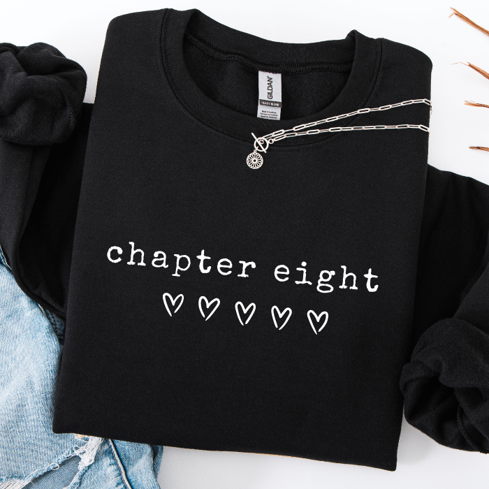 A black crewneck sweatshirt with 'chapter eight' and four outlined hearts printed in white on the front, styled flat-lay with a silver chain necklace and light blue denim.