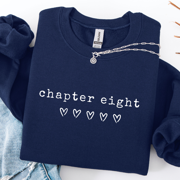A navy blue sweatshirt with 'chapter eight' and five outlined hearts printed in white on the front, styled flat-lay with light blue jeans and a silver chain necklace on a white background.