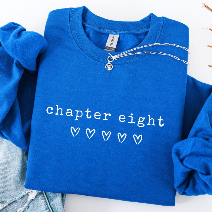 A folded royal blue sweatshirt with rolled sleeves, featuring the words 'chapter eight' and five outlined hearts printed in white on the front. A silver chain necklace is draped across the chest, and light-wash denim jeans are partially visible at the bottom left.