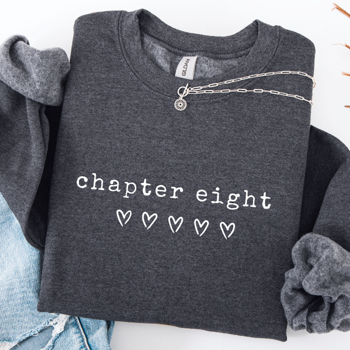 A folded dark heather gray sweatshirt with the words 'chapter eight' and five small heart icons printed in white on the front. A silver chain necklace is draped across the collar area, and the sweatshirt is styled with light blue jeans on a white background.