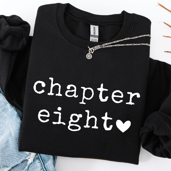 A black crewneck sweatshirt with 'chapter eight' printed in white typewriter-style text and a white heart, styled flat-lay with a silver chain necklace and light blue denim.