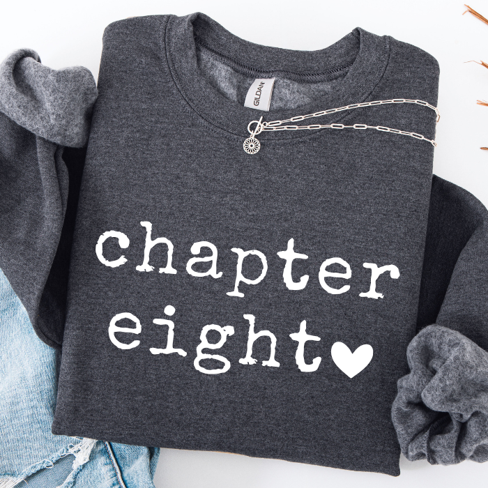 A dark heather gray sweatshirt with 'chapter eight' printed in white across the front, styled flat-lay with rolled sleeves, a silver chain necklace, and light blue jeans on a white background.