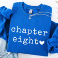A folded royal blue sweatshirt with rolled sleeves, featuring the words 'chapter eight' in white distressed lettering and a white heart. A silver chain necklace is draped across the collar, and light-wash denim jeans are partially visible underneath on a white background.