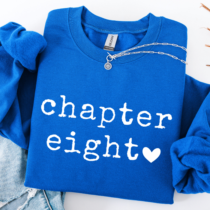 A folded royal blue sweatshirt with rolled sleeves, featuring the words 'chapter eight' in white distressed lettering and a white heart. A silver chain necklace is draped across the collar, and light-wash denim jeans are partially visible underneath on a white background.