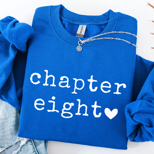 A folded royal blue sweatshirt with rolled sleeves, featuring the words 'chapter eight' in white distressed lettering and a white heart. A silver chain necklace is draped across the collar, and light-wash denim jeans are partially visible underneath on a white background.