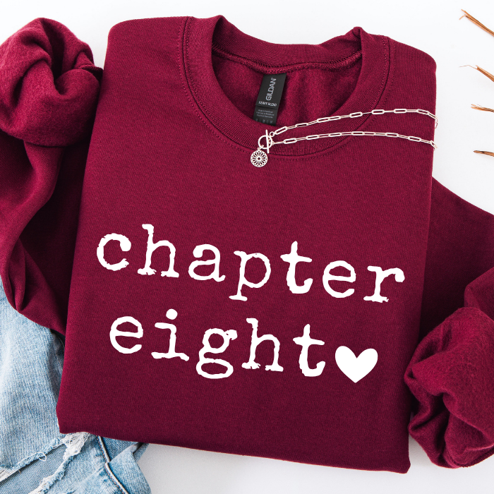 A folded burgundy sweatshirt with the words 'chapter eight' and a small heart printed in white on the front, styled with a silver chain necklace and light blue jeans on a white background.