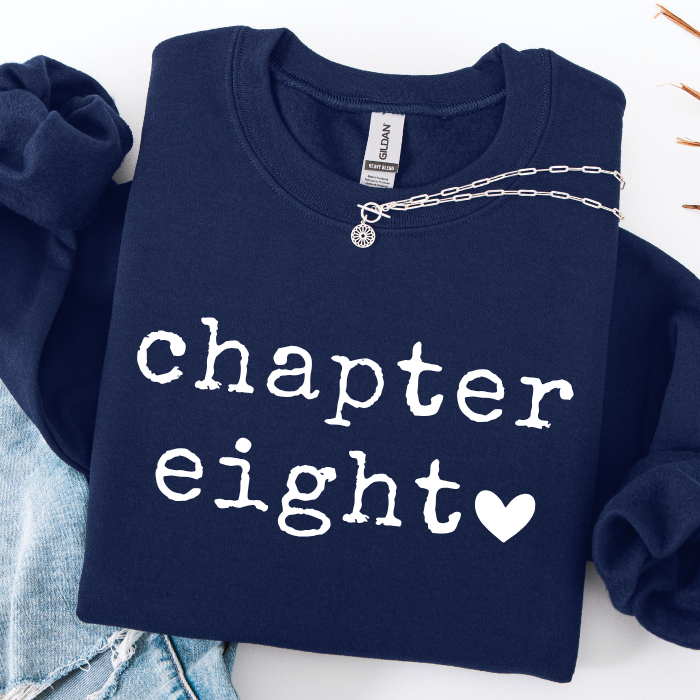 A navy blue sweatshirt with 'chapter eight' and a heart printed in white on the front, styled flat-lay with light blue jeans and a silver chain necklace on a white background.