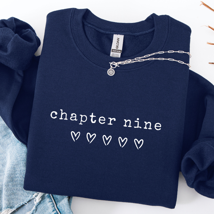 A navy blue sweatshirt with 'chapter nine' and five outlined hearts printed in white on the front, styled flat-lay with a silver chain necklace and light blue denim visible around it.