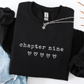 A folded black crewneck sweatshirt with 'chapter nine' and five outlined hearts printed in white on the front, styled with a silver chain necklace and light blue denim on a white background.
