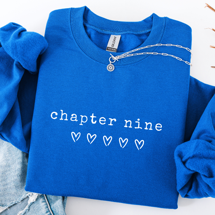 A folded royal blue sweatshirt with the words 'chapter nine' and five small heart icons printed in white across the front. The sweatshirt is styled on a white surface with rolled sleeves and a silver chain necklace draped over the collar.
