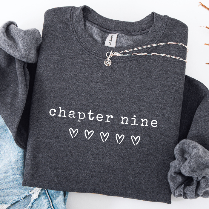 A dark heather gray sweatshirt with 'chapter nine' and five hearts printed in white on the front, styled flat-lay with rolled sleeves, a silver chain necklace, and light blue jeans on a white background.