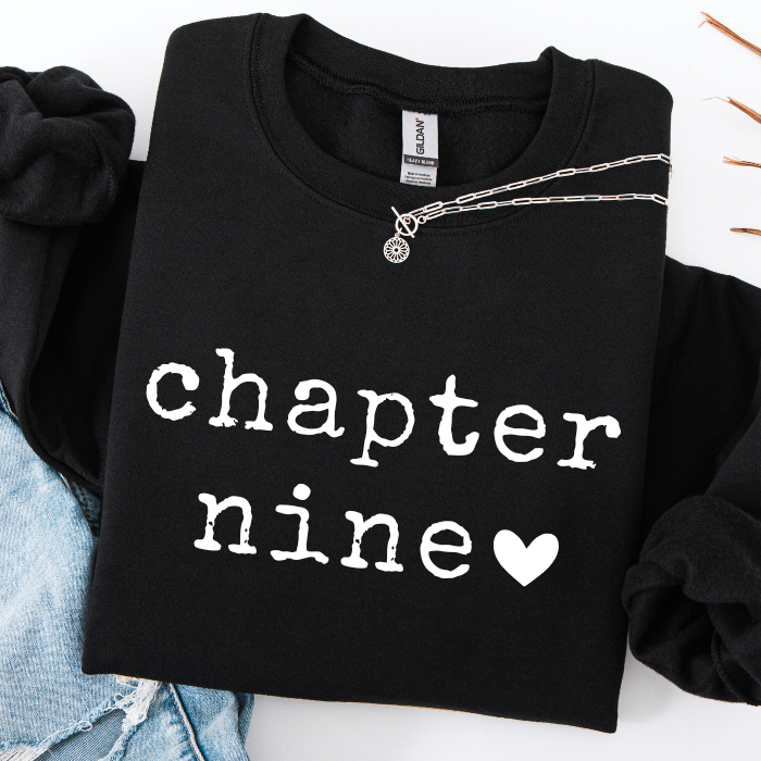 A black crewneck sweatshirt with 'chapter nine' and a heart printed in white on the front, styled flat-lay with a silver chain necklace and light blue denim.