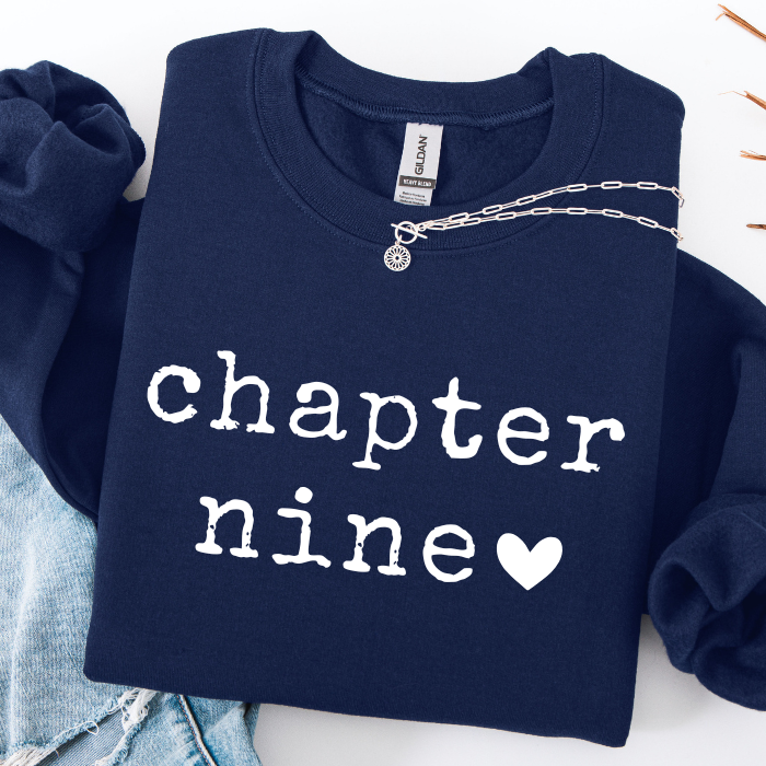 A navy blue sweatshirt with 'chapter nine' and a heart printed in white on the front, styled flat-lay with light blue jeans and a silver chain necklace on a white background.