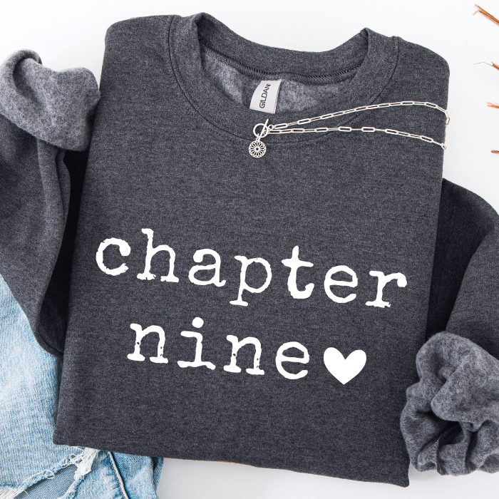 A dark heather gray sweatshirt with 'chapter nine' printed in white lowercase letters and a white heart, styled flat-lay with rolled sleeves, a silver chain necklace, and light blue jeans on a white background.