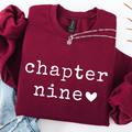 A folded burgundy sweatshirt with 'chapter nine' and a heart printed in white on the front, styled with a silver chain necklace and light blue jeans on a white background.