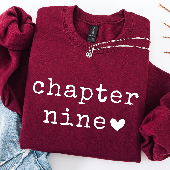 A folded burgundy sweatshirt with 'chapter nine' and a heart printed in white on the front, styled with a silver chain necklace and light blue jeans on a white background.