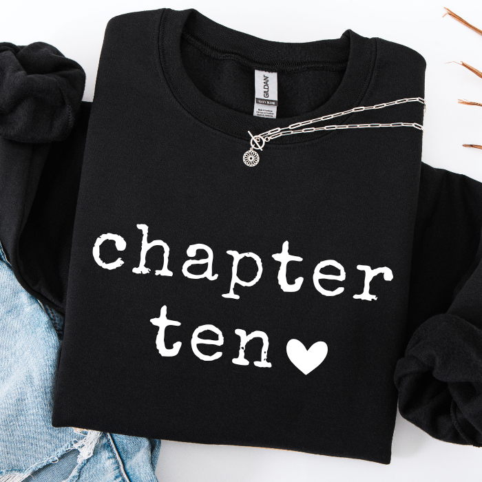 A folded black sweatshirt with the words 'chapter ten' and a white heart printed on the front, styled with a silver chain necklace and light blue denim on a white background.