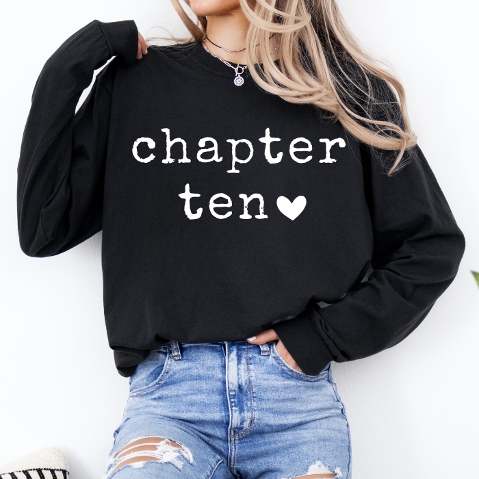 A woman wearing a black long-sleeve sweatshirt with 'chapter ten' printed in white text and a small white heart on the front, paired with distressed blue jeans.
