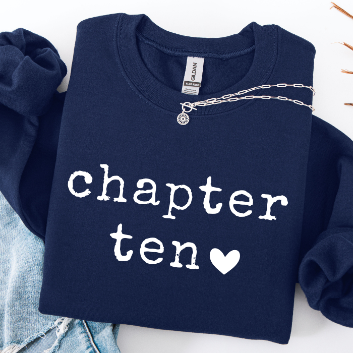 A navy blue sweatshirt with 'chapter ten' and a heart printed in white across the front, styled flat-lay with a silver chain necklace and light blue denim folded around it on a white background.