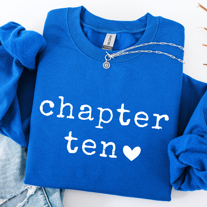 A folded royal blue sweatshirt with rolled sleeves, featuring the words 'chapter ten' in white script and a white heart. A silver chain necklace is draped across the chest, and light-wash denim jeans are partially visible at the bottom left.