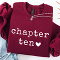 A folded burgundy sweatshirt with 'chapter ten' printed in white across the front, styled with a silver chain necklace and light blue denim on a white background.