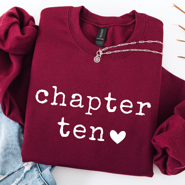 A folded burgundy sweatshirt with 'chapter ten' printed in white across the front, styled with a silver chain necklace and light blue denim on a white background.