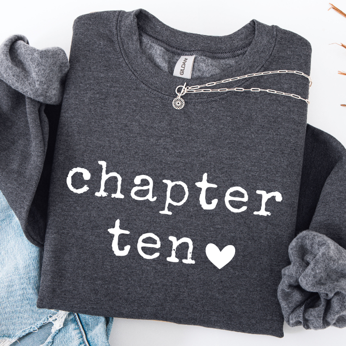 A dark heather gray sweatshirt with 'chapter ten' printed in white typewriter-style text and a small white heart, styled flat-lay with rolled sleeves, a silver chain necklace, and light blue jeans on a white background.