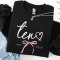A folded black sweatshirt with white script text reading 'ten' and a heart, plus a pink bow graphic and 'est 2016' printed on the front. A silver necklace is draped across the collar, and the sweatshirt is styled with light blue denim on a white background.