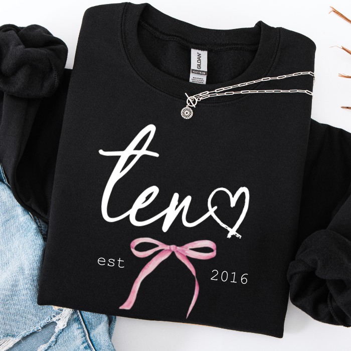 A folded black sweatshirt with white script text reading 'ten' and a heart, plus a pink bow graphic and 'est 2016' printed on the front. A silver necklace is draped across the collar, and the sweatshirt is styled with light blue denim on a white background.