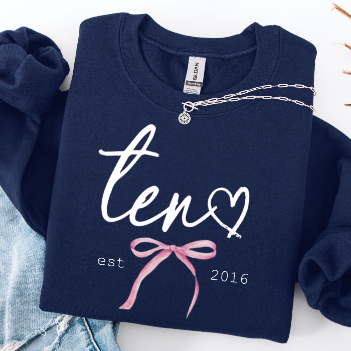 A navy blue sweatshirt laid flat on a white surface, featuring a white script 'ten' with a heart and key graphic, plus 'est 2016' and a pink bow design. A silver chain necklace is draped across the chest, and light blue denim is partially visible at the lower left.