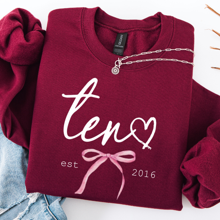 A folded burgundy sweatshirt with a white script 'ten' and heart graphic, plus a pink bow and 'est 2016' print, styled on a white background with a silver necklace draped across the collar and light blue jeans partially visible at the side.